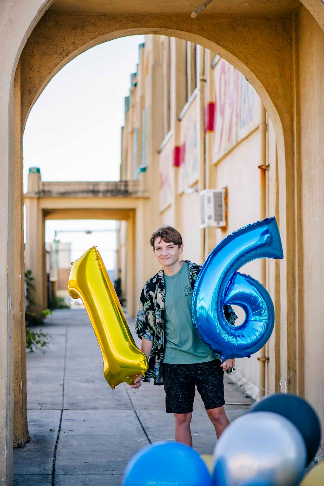 Teen boy holding gold number 1 and blue number 6 balloons celebrating his 16th birthday in an outdoor alleyway.