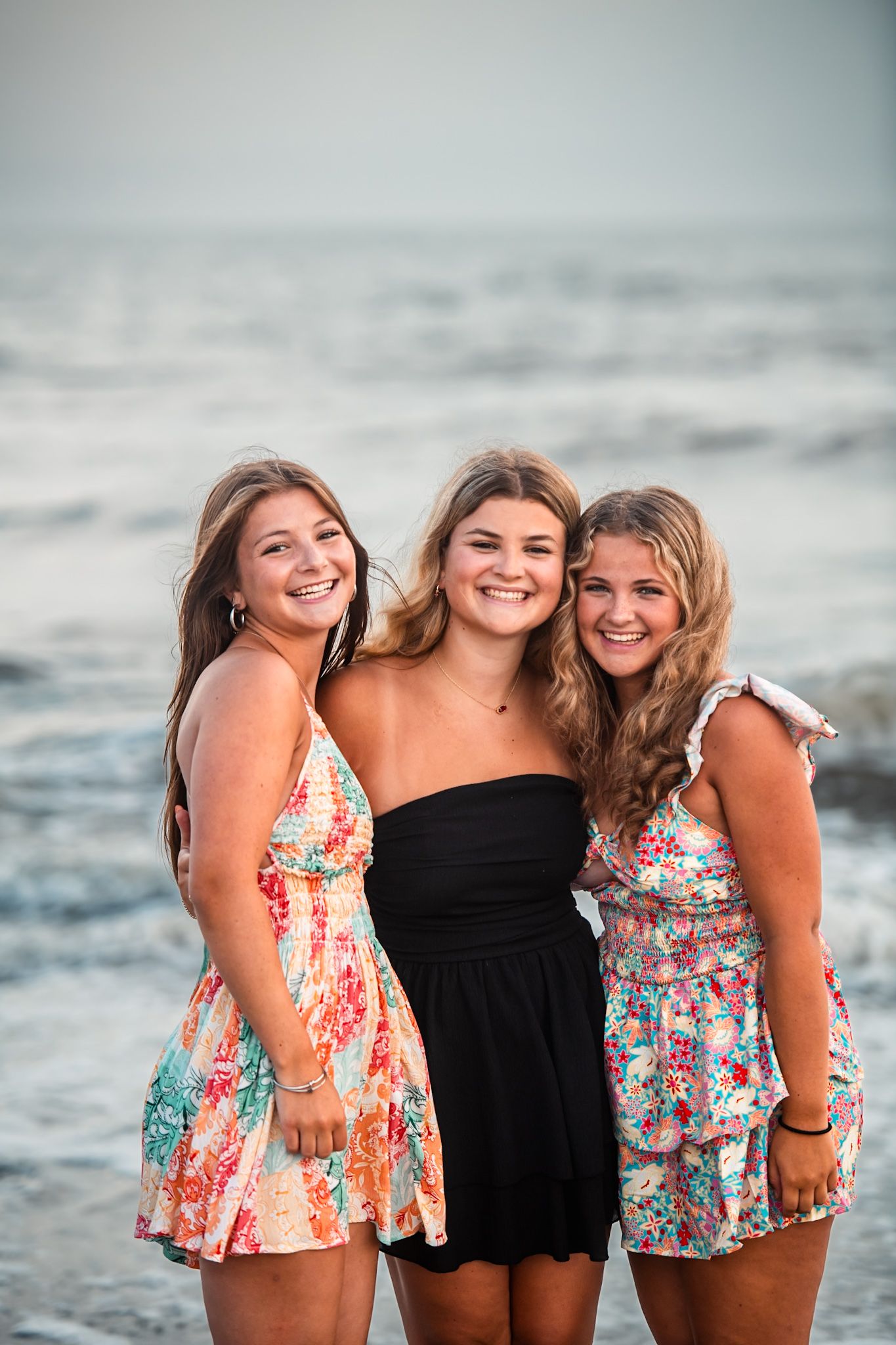 Three smiling teenage girls in summer dresses pose together on a beach with ocean waves in the background.