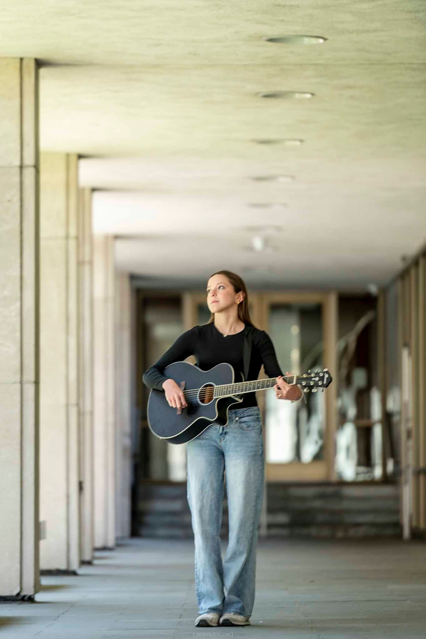 Young woman in black top and jeans playing acoustic guitar while walking through an urban colonnade.