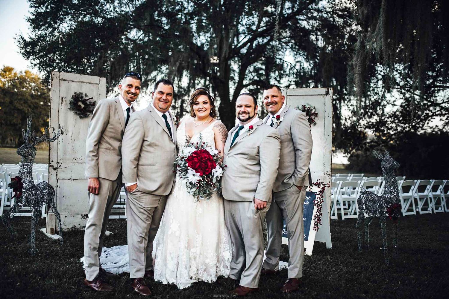 Bride in white lace gown poses with groomsmen in tan suits holding red bouquet at outdoor wedding.