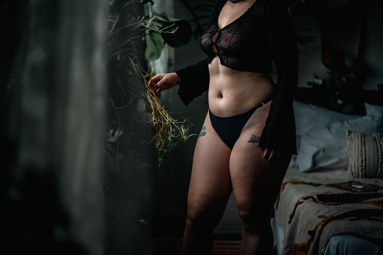 Person in black lingerie holding dried herbs in a moody, dimly lit bedroom setting.