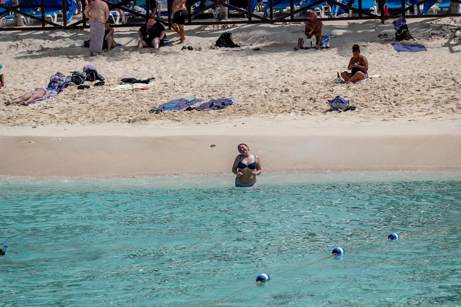 Tourists relaxing on a white sandy Caribbean beach with crystal clear turquoise water and blue buoy markers.