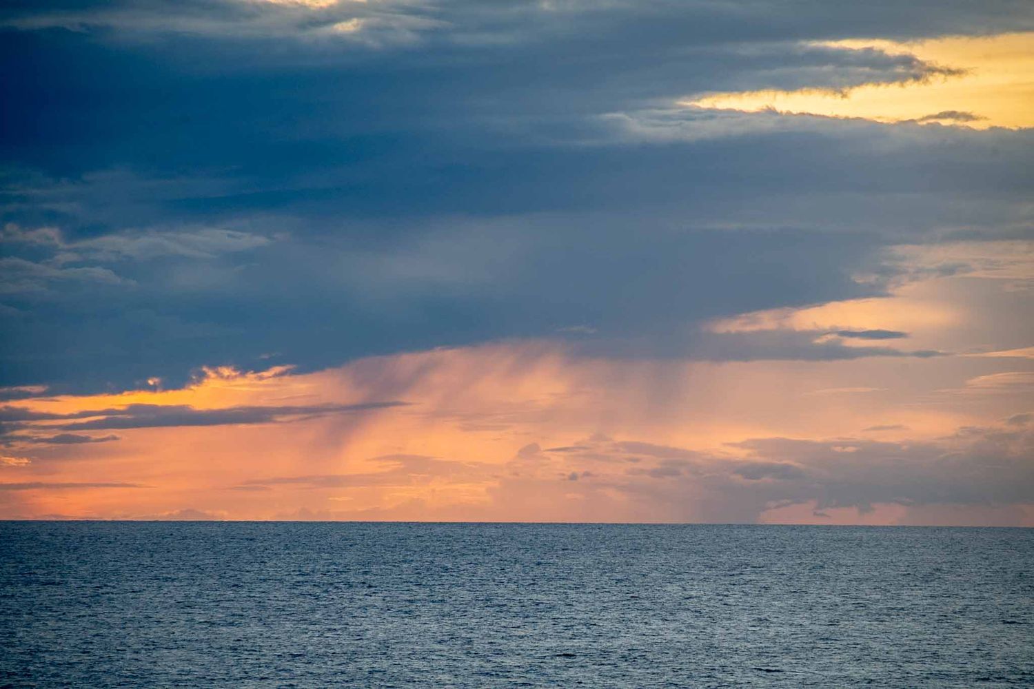 Dramatic sunset sky with orange rays breaking through dark clouds over a calm ocean horizon.
