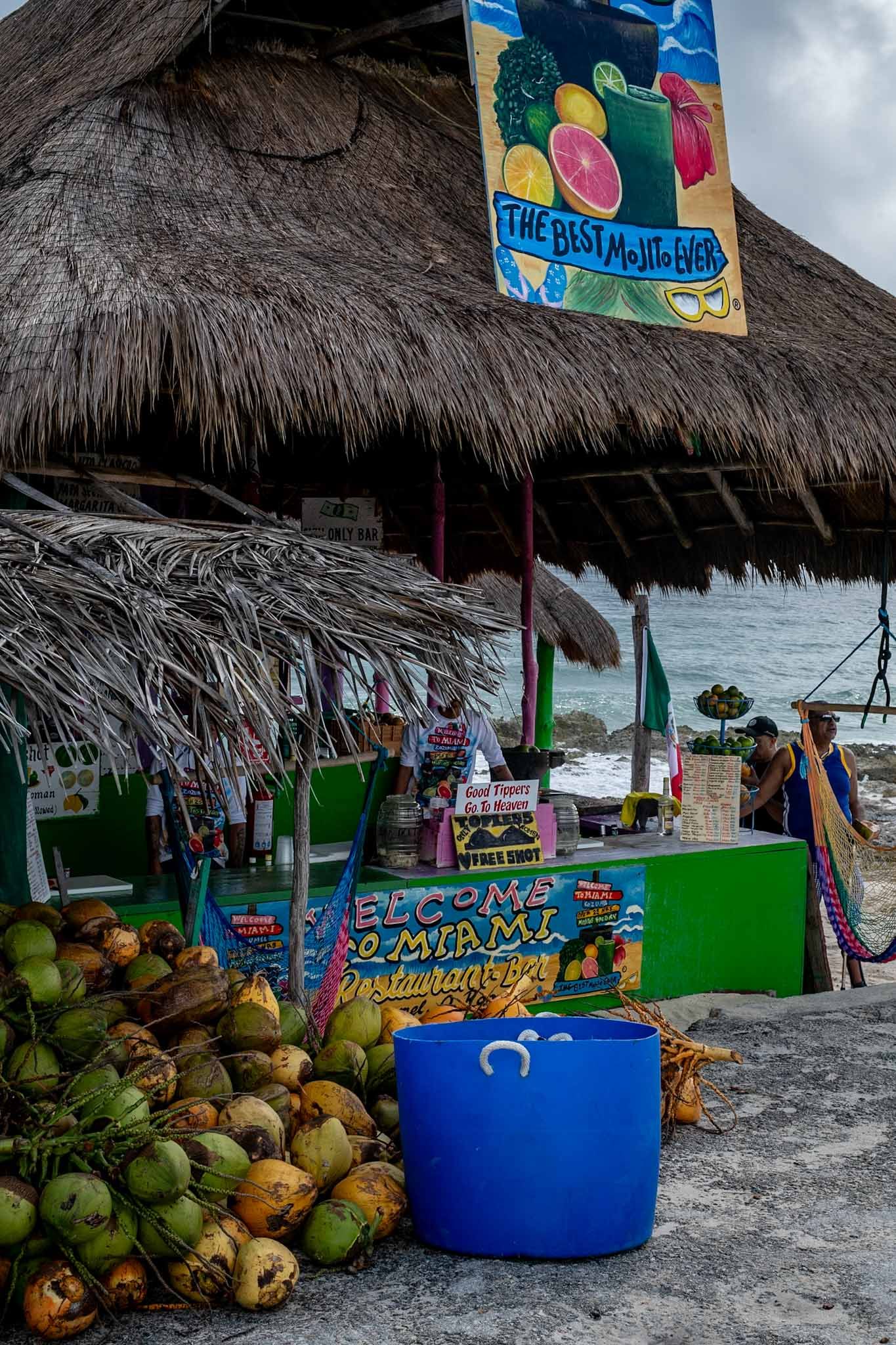 Beach vendor stall with thatched roof selling fresh coconuts and tropical drinks on a cloudy day.