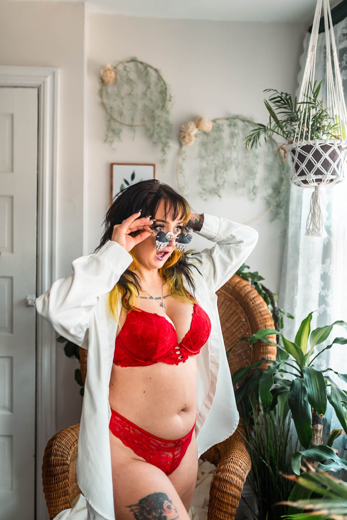 Woman in red lingerie and white robe adjusting sunglasses in a plant-filled bright room.