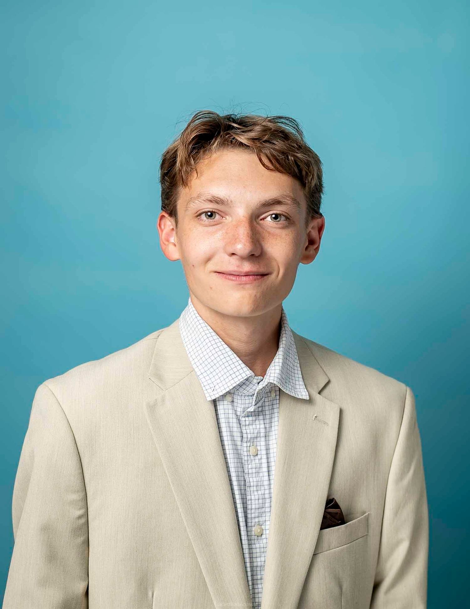 Young man smiling in beige blazer and checkered shirt against blue background, professional headshot.