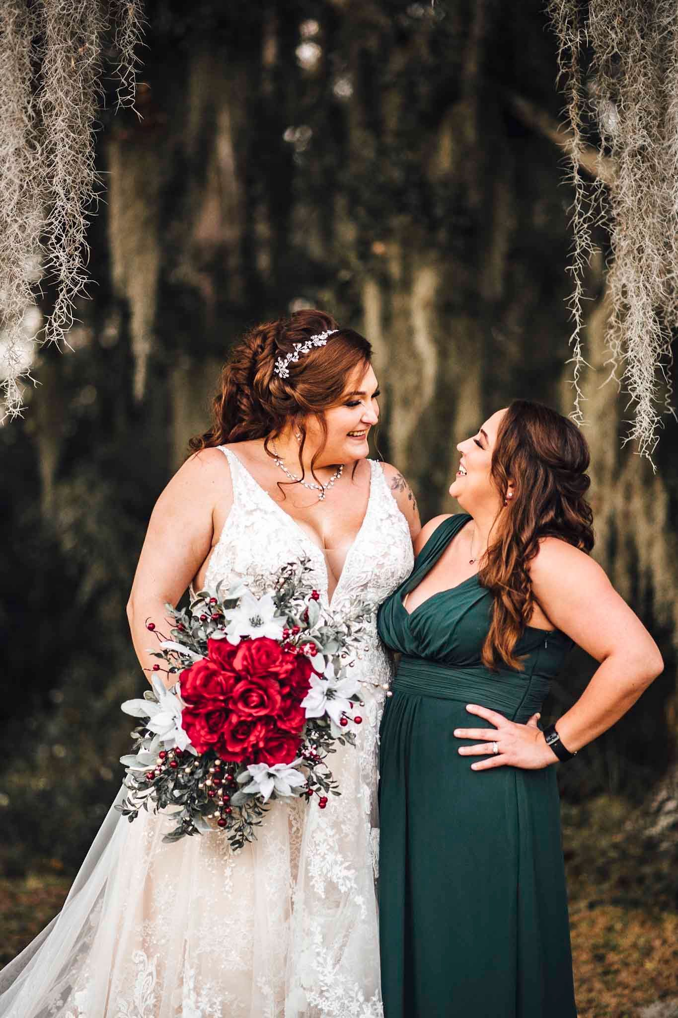 Bride in lace gown holds red rose bouquet beside bridesmaid in emerald dress under Spanish moss trees.