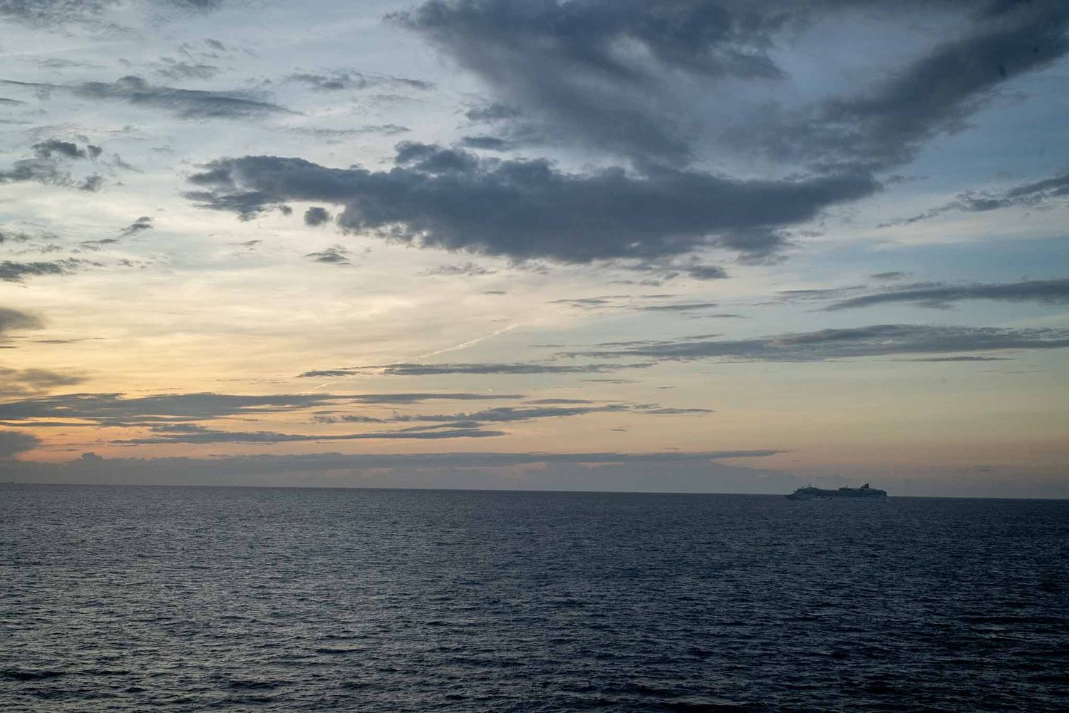 Dramatic sunset over calm ocean waters with dark clouds and distant ships on the horizon.