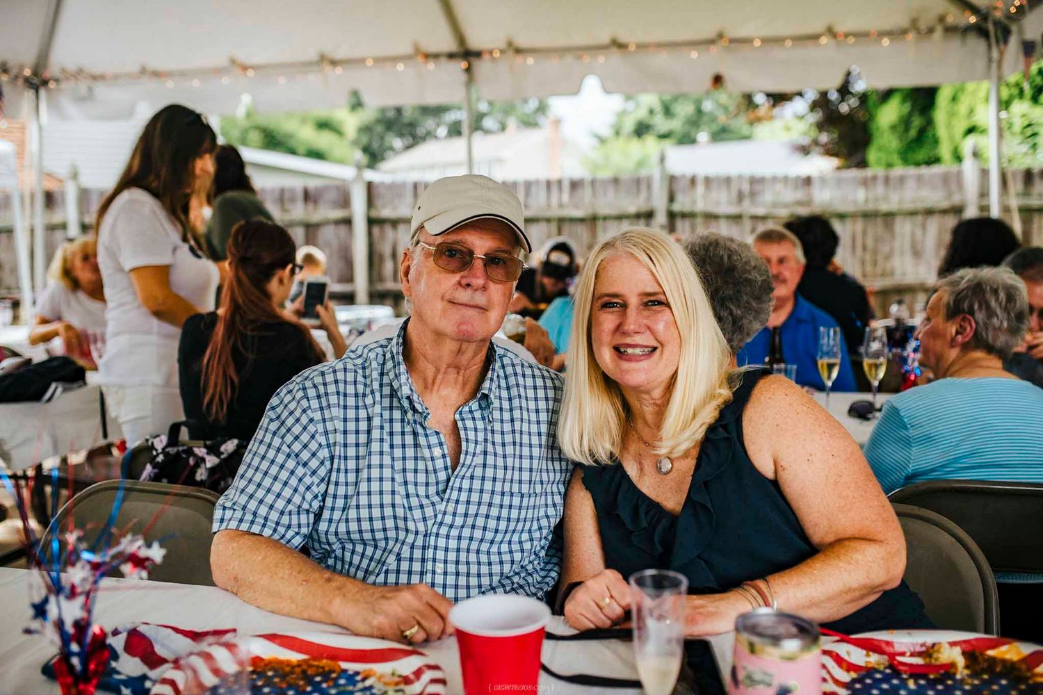 Smiling couple poses together at a patriotic outdoor party under a white tent with festive decorations.