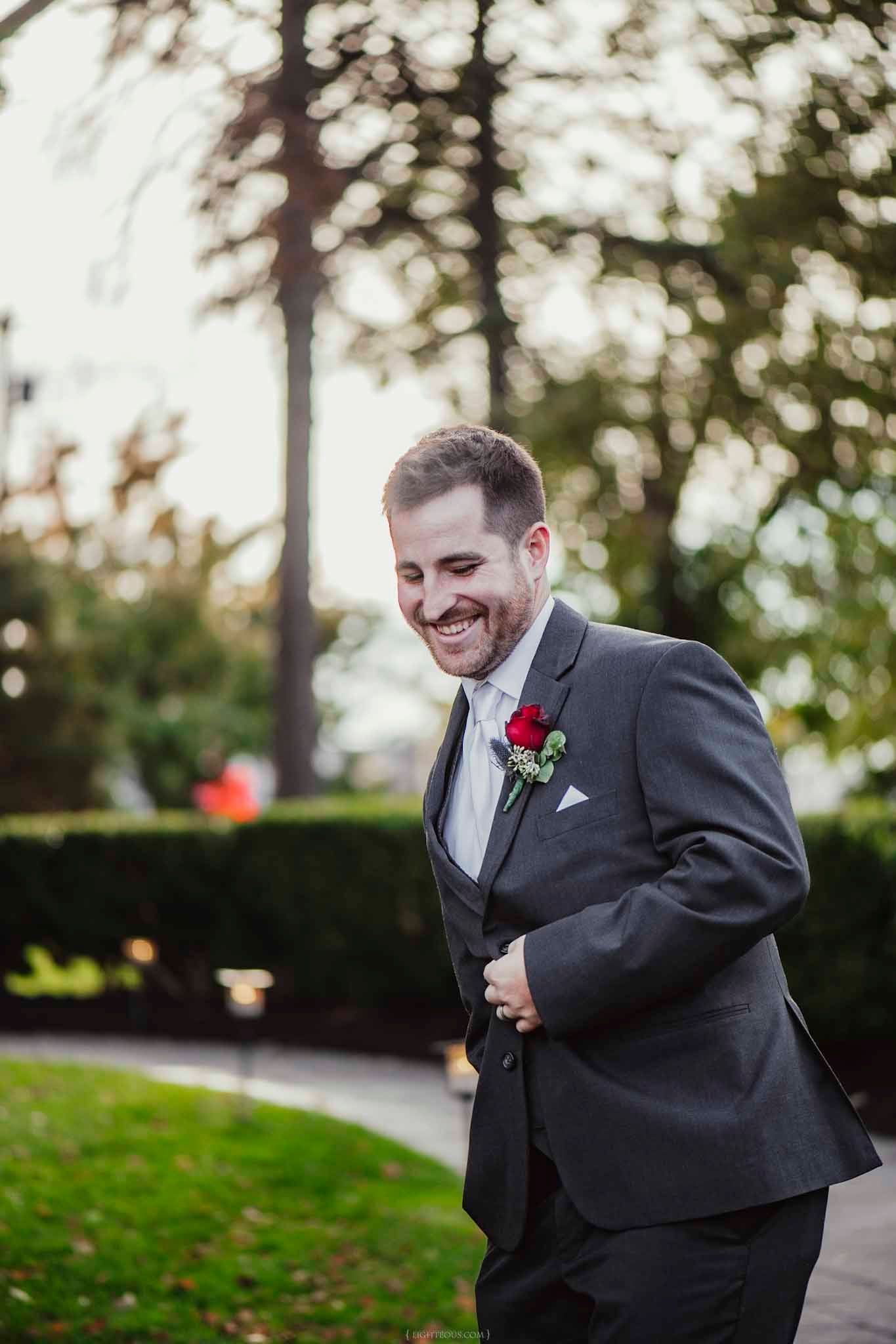 Smiling groom in gray suit with red rose boutonniere adjusting jacket outdoors at wedding ceremony.