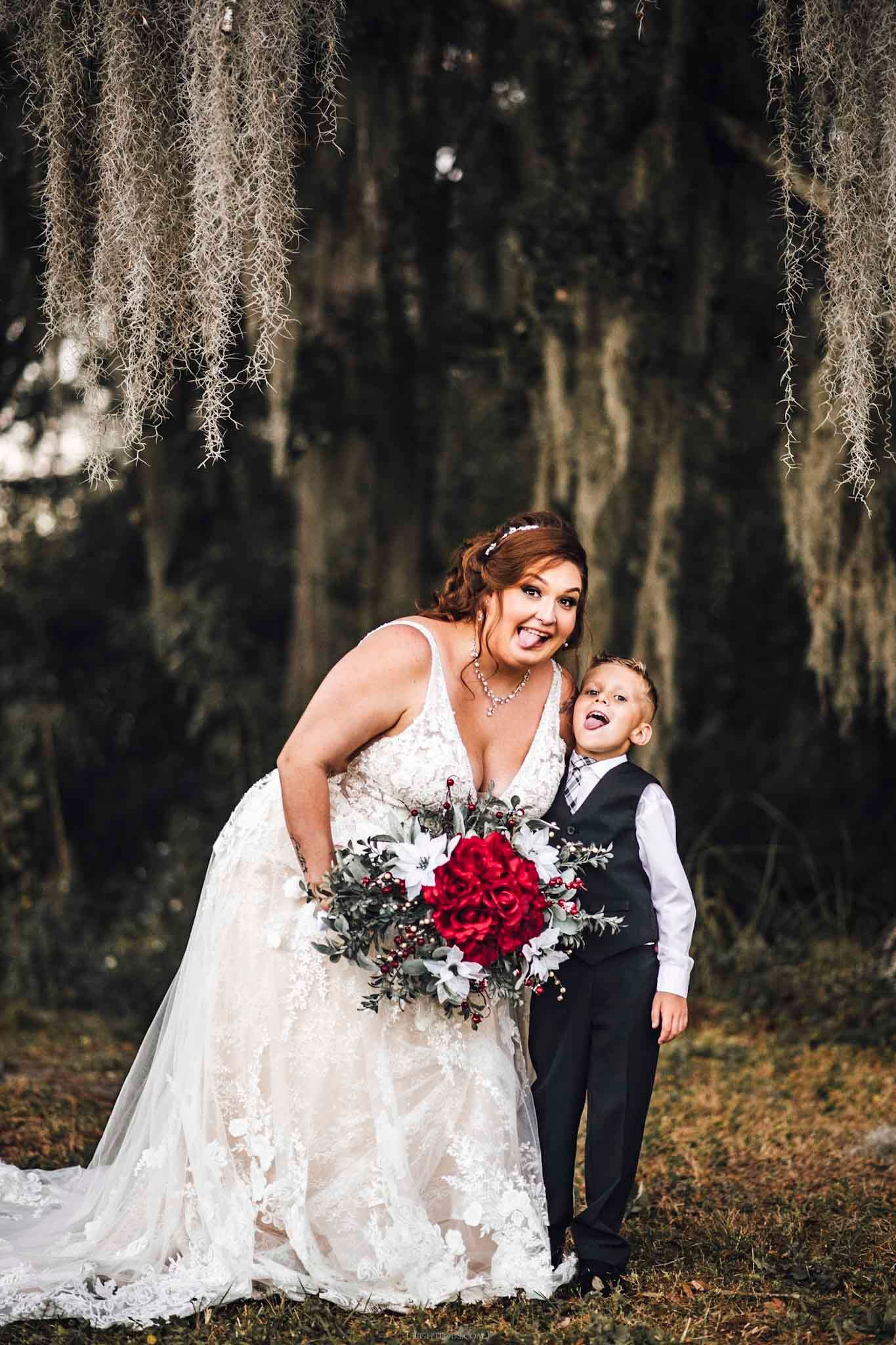Bride in lace gown holding red bouquet poses with young boy in suit under Spanish moss trees.