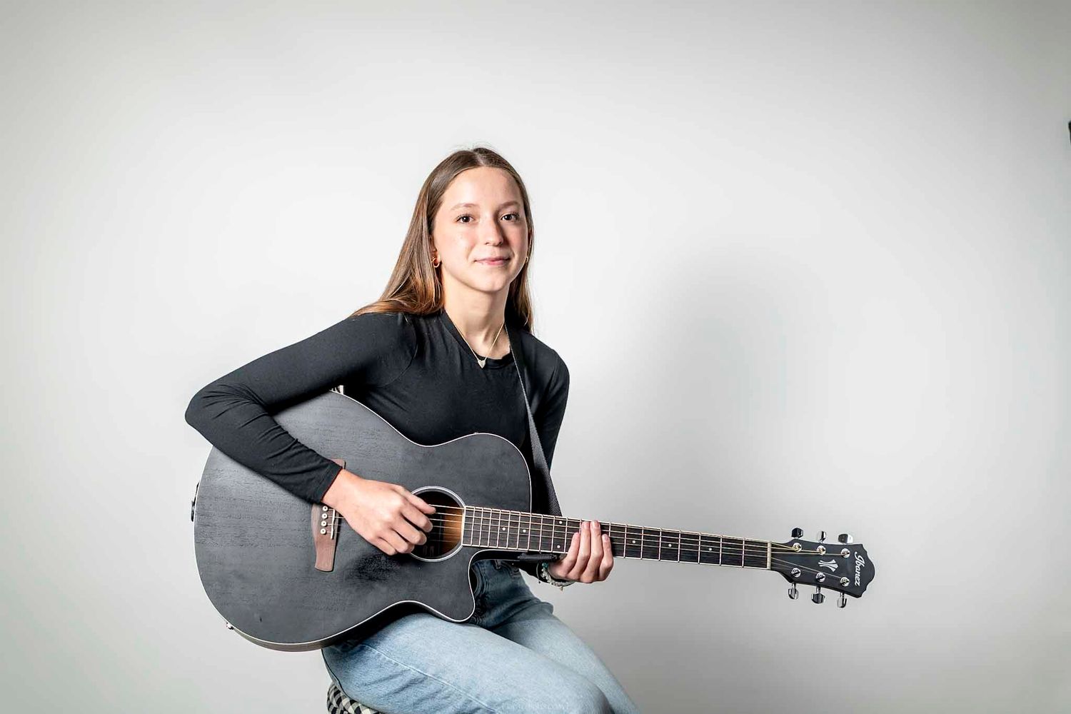 Young woman sitting and playing a black acoustic guitar, wearing a black top and jeans, against a white background.