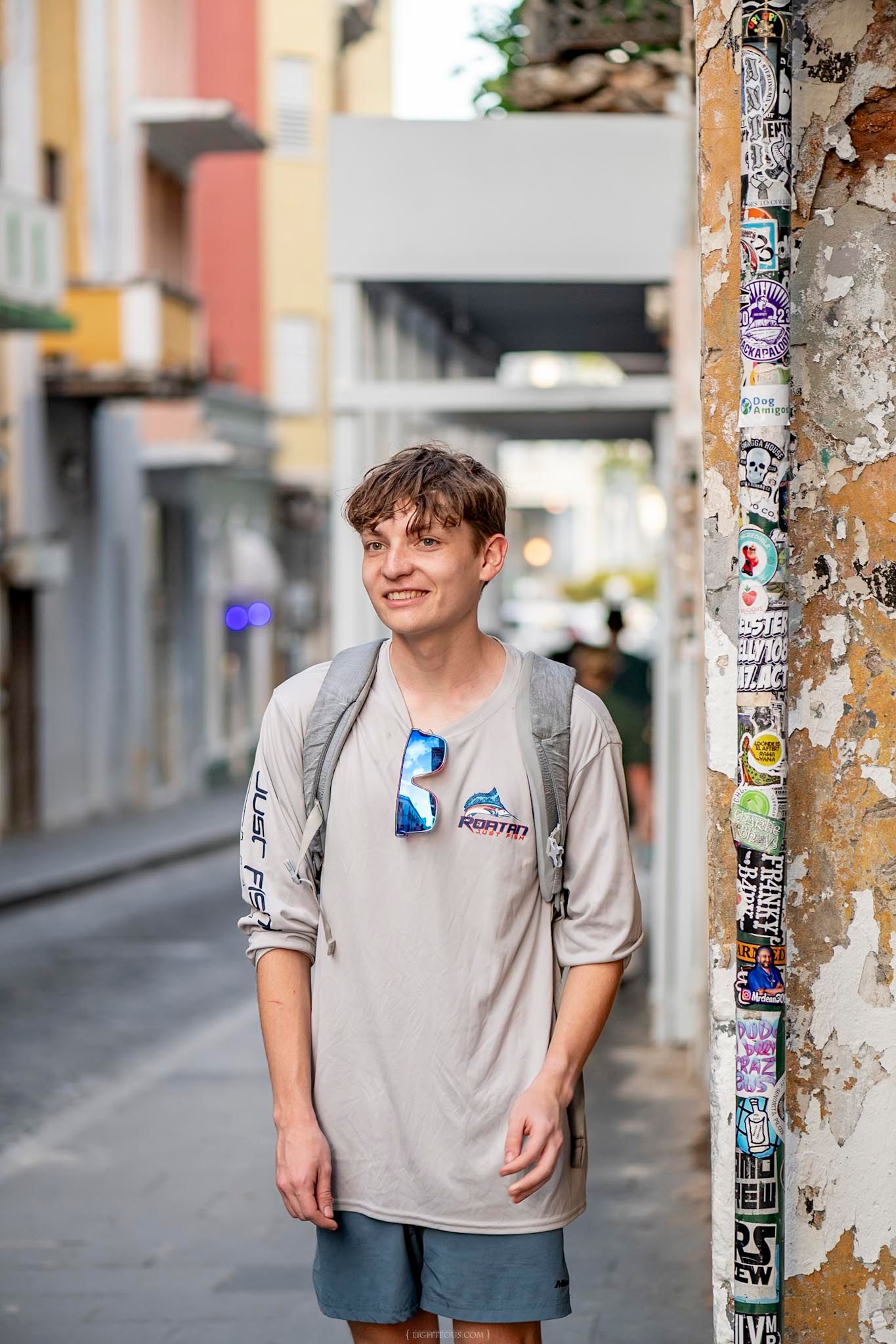 Young man in gray t-shirt and backpack smiling on a colorful European street beside a sticker-covered pillar.
