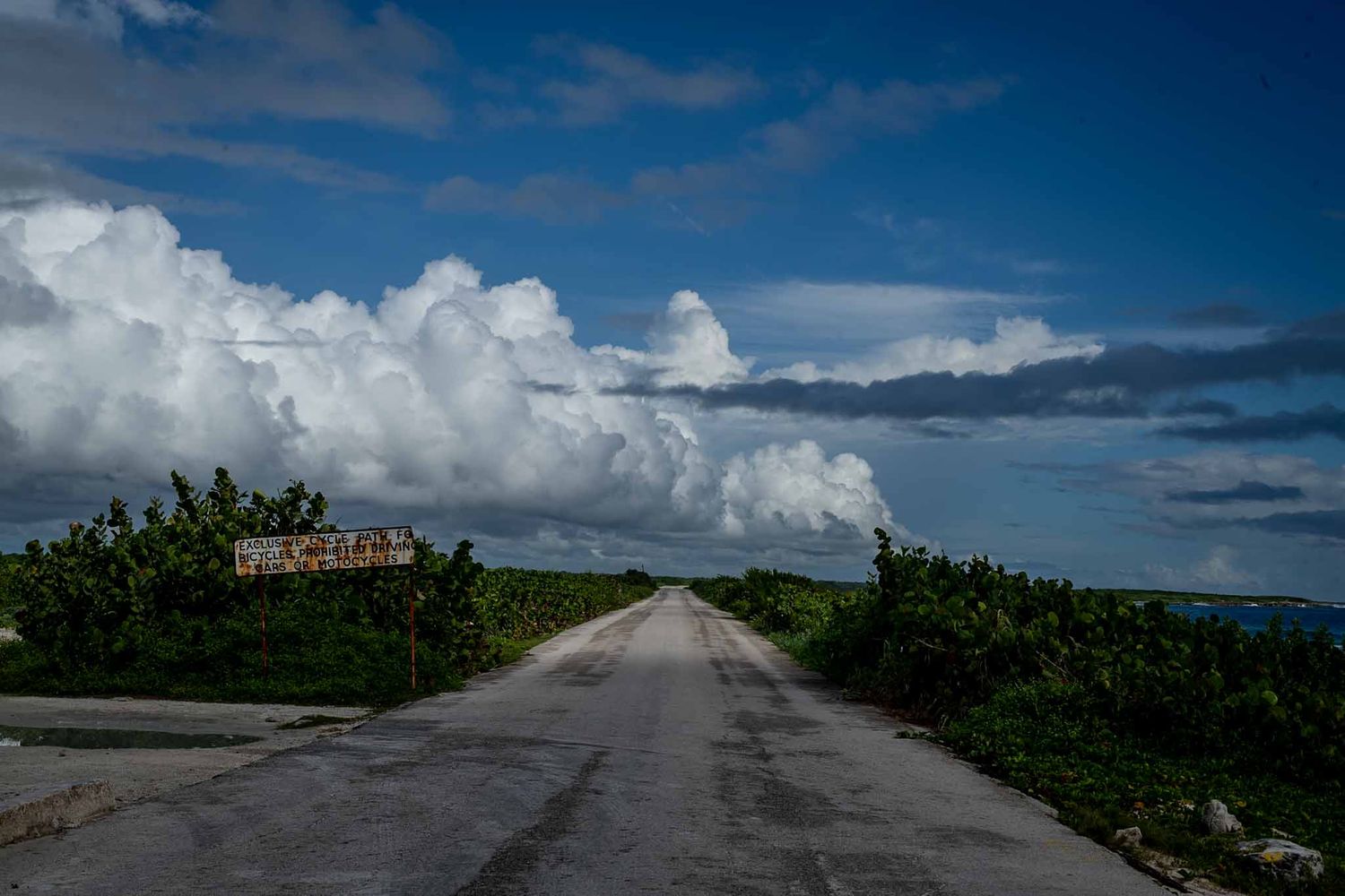 Empty road stretches into distance under dramatic cloudy sky with lush vegetation on both sides.