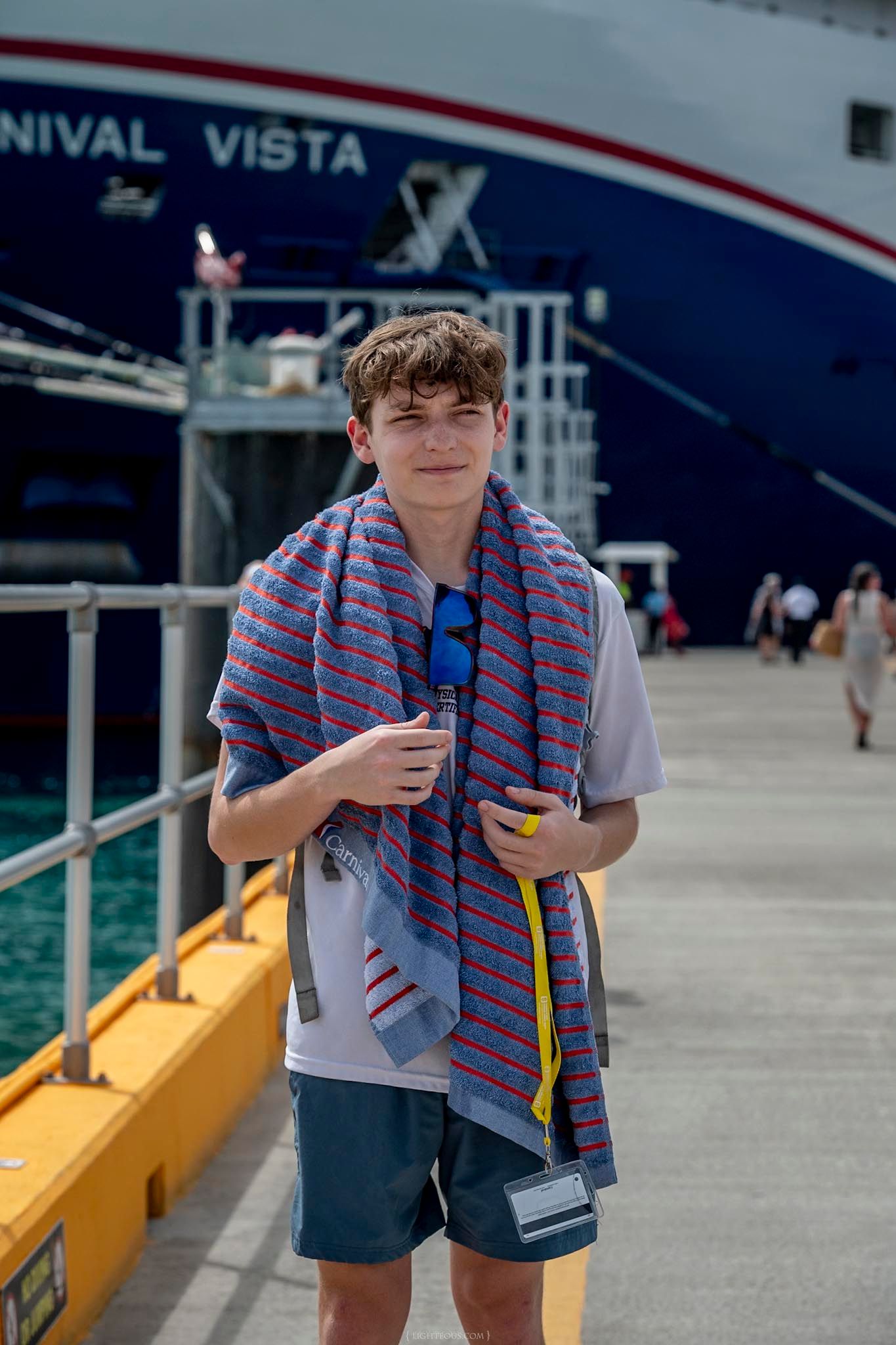 Teen boy with striped towel stands on dock in front of Carnival Vista cruise ship on sunny day.