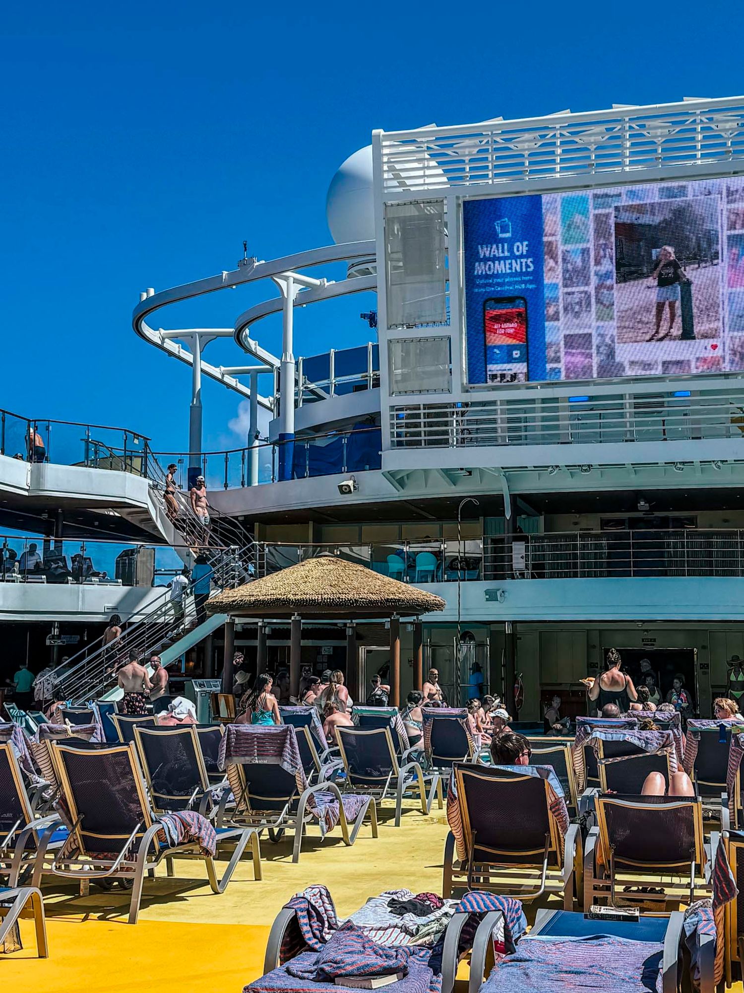 Passengers relaxing on sun deck chairs of cruise ship with water slide and large LED screen display.