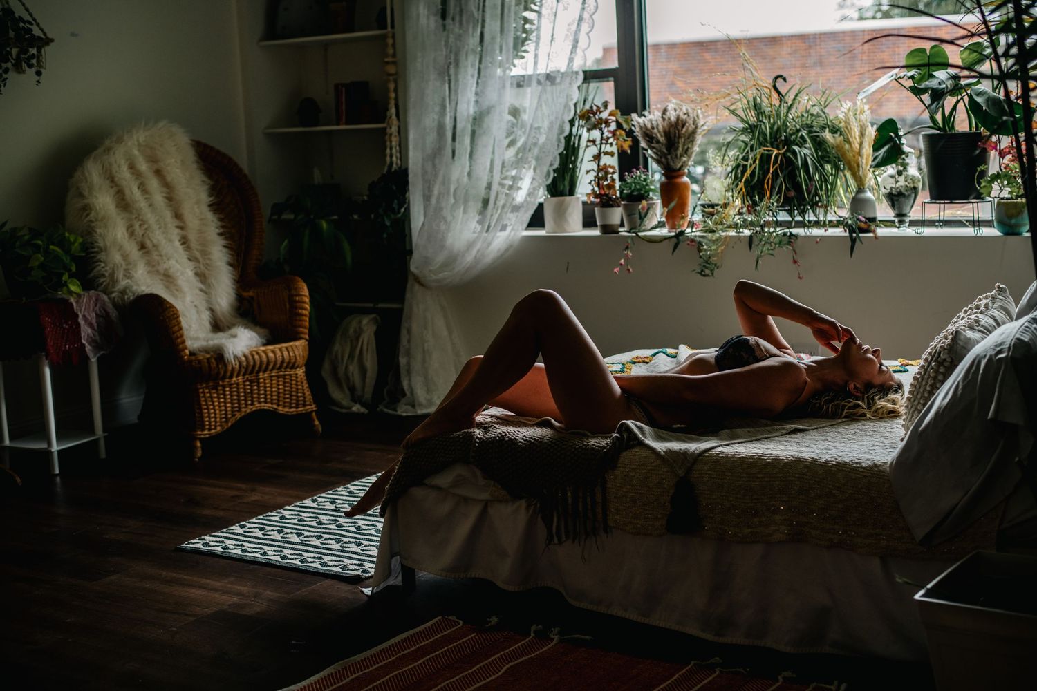 A woman relaxes on a bed in a cozy, plant-filled room with warm natural light streaming through sheer curtains.