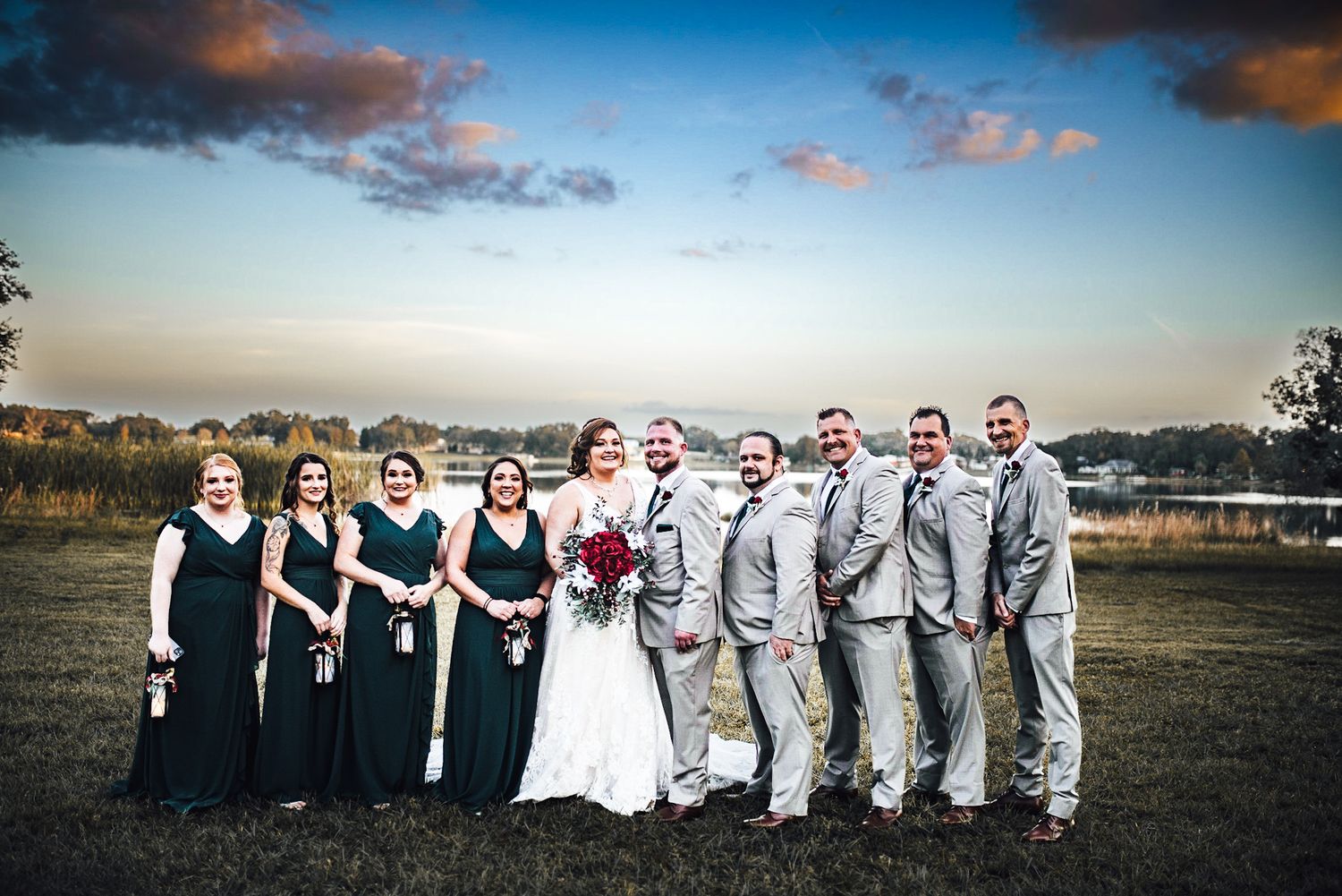 Wedding party posing outdoors at sunset, bridesmaids in green gowns and groomsmen in gray suits flanking bride and groom.