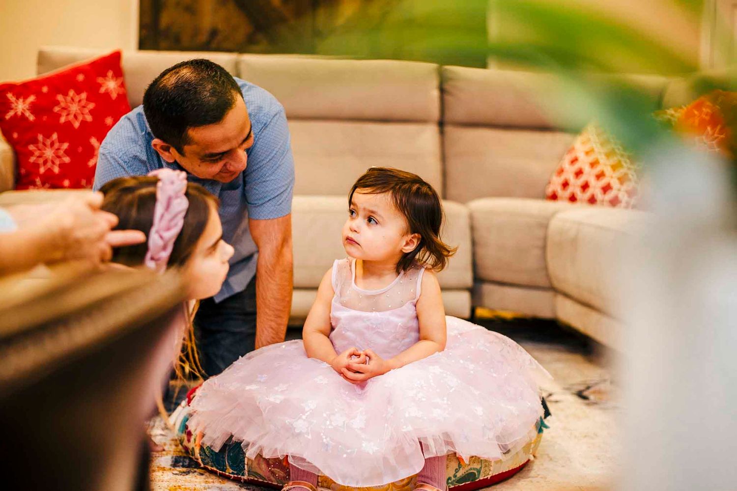 A little girl in a pink tutu dress sits on a cushion as a man leans down to interact with her indoors.