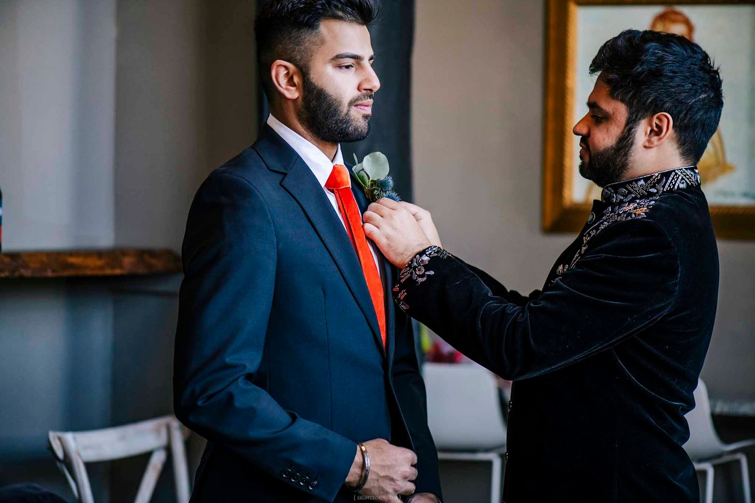 A man in a navy suit with red tie gets a boutonniere pinned by another man in a black velvet jacket.