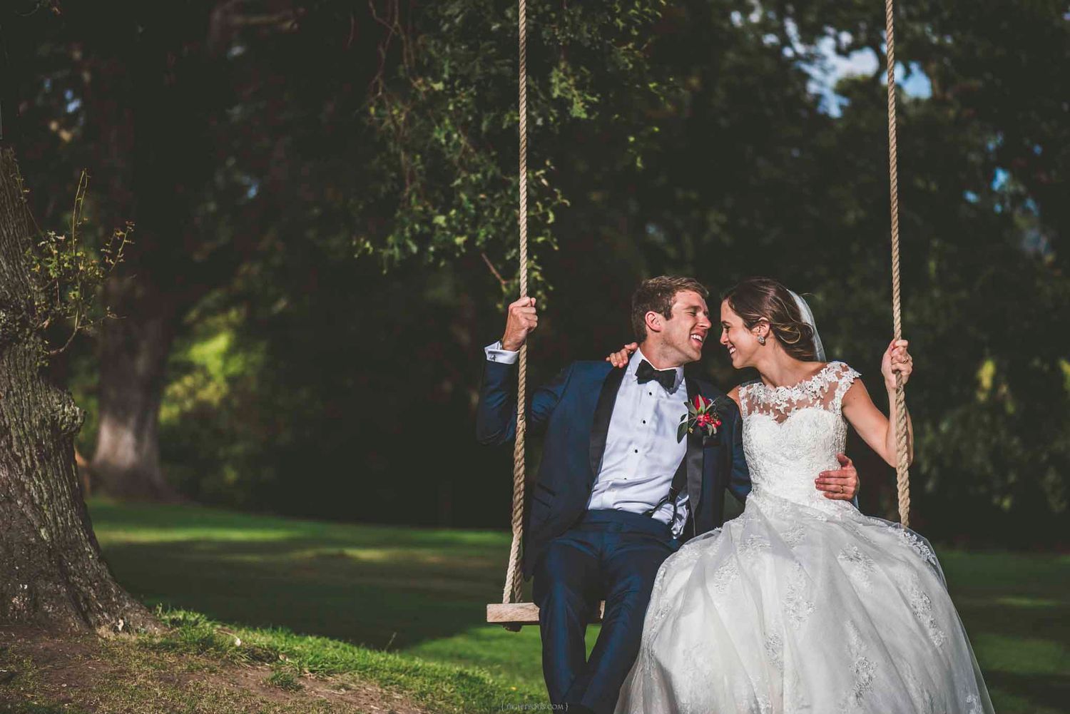 A bride and groom share a romantic moment on a rope swing under a large tree in a lush green garden.