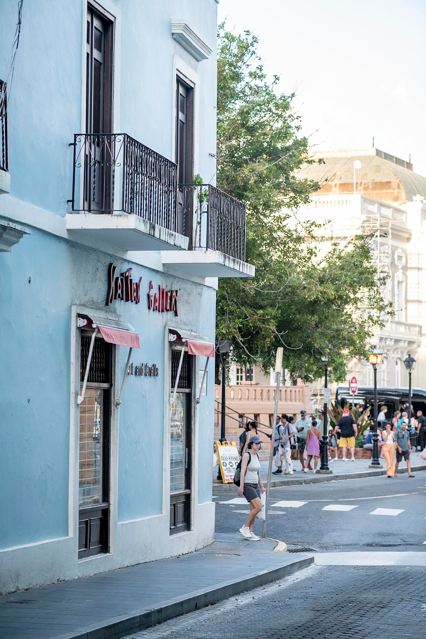 Pedestrians walk past a light blue corner building with a gallery shop in a sunny colonial street scene.