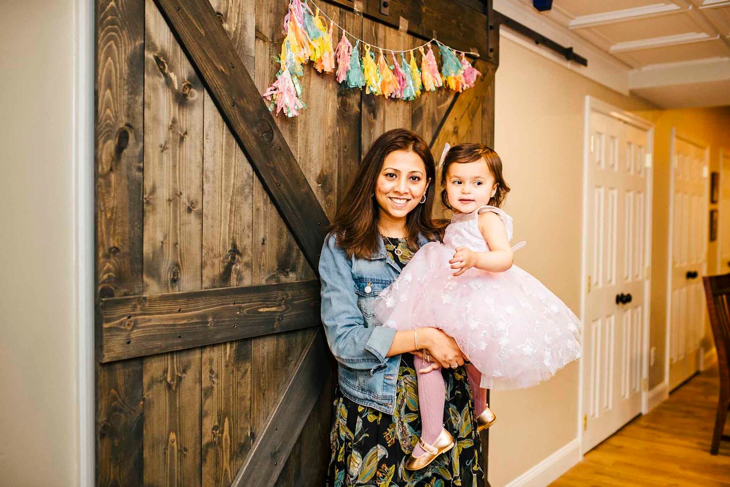 Woman in denim jacket holds toddler girl in pink tutu dress at colorful birthday party with barn door backdrop.