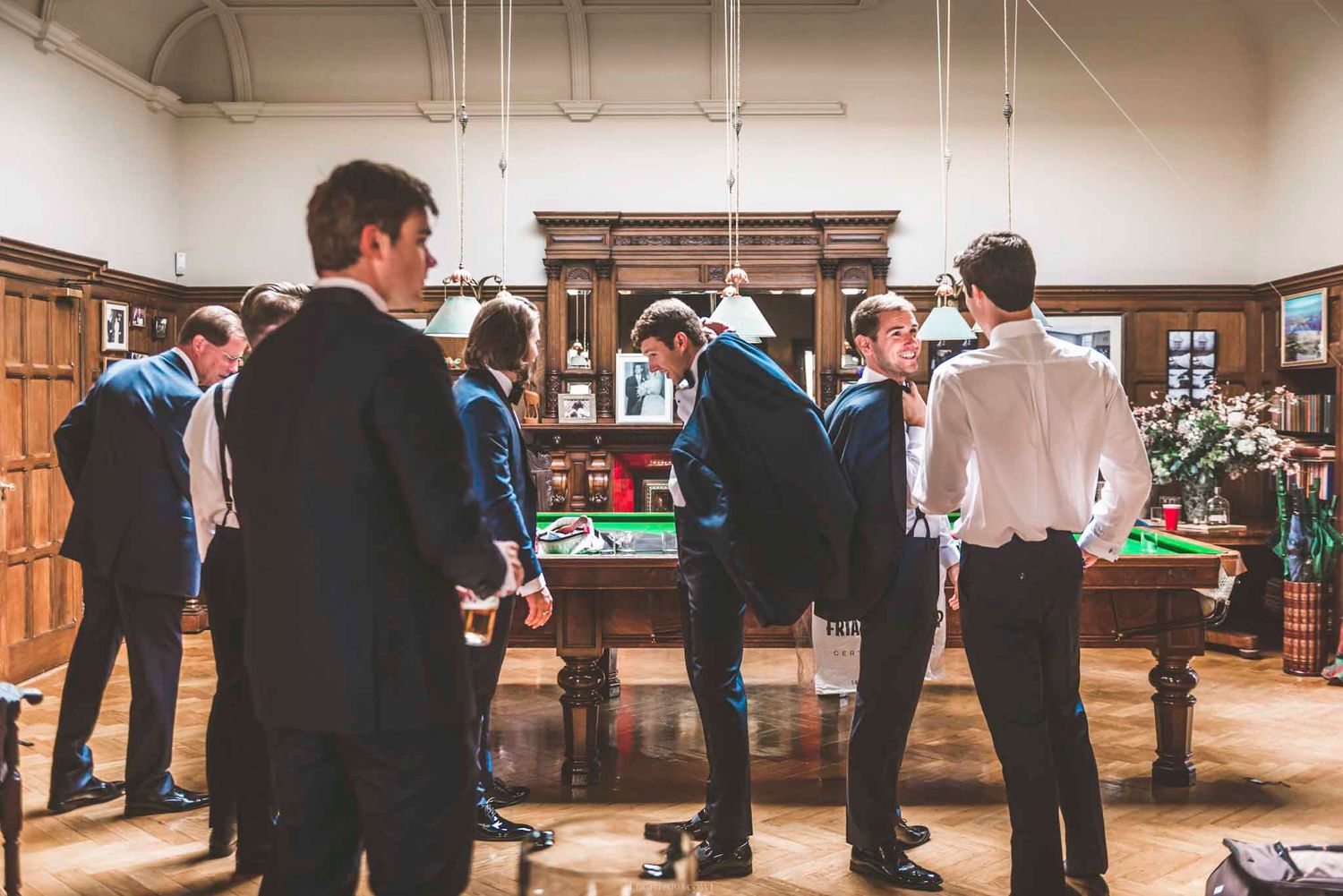 Wedding guests in suits gathered around a billiard table in an elegant wood-paneled room.