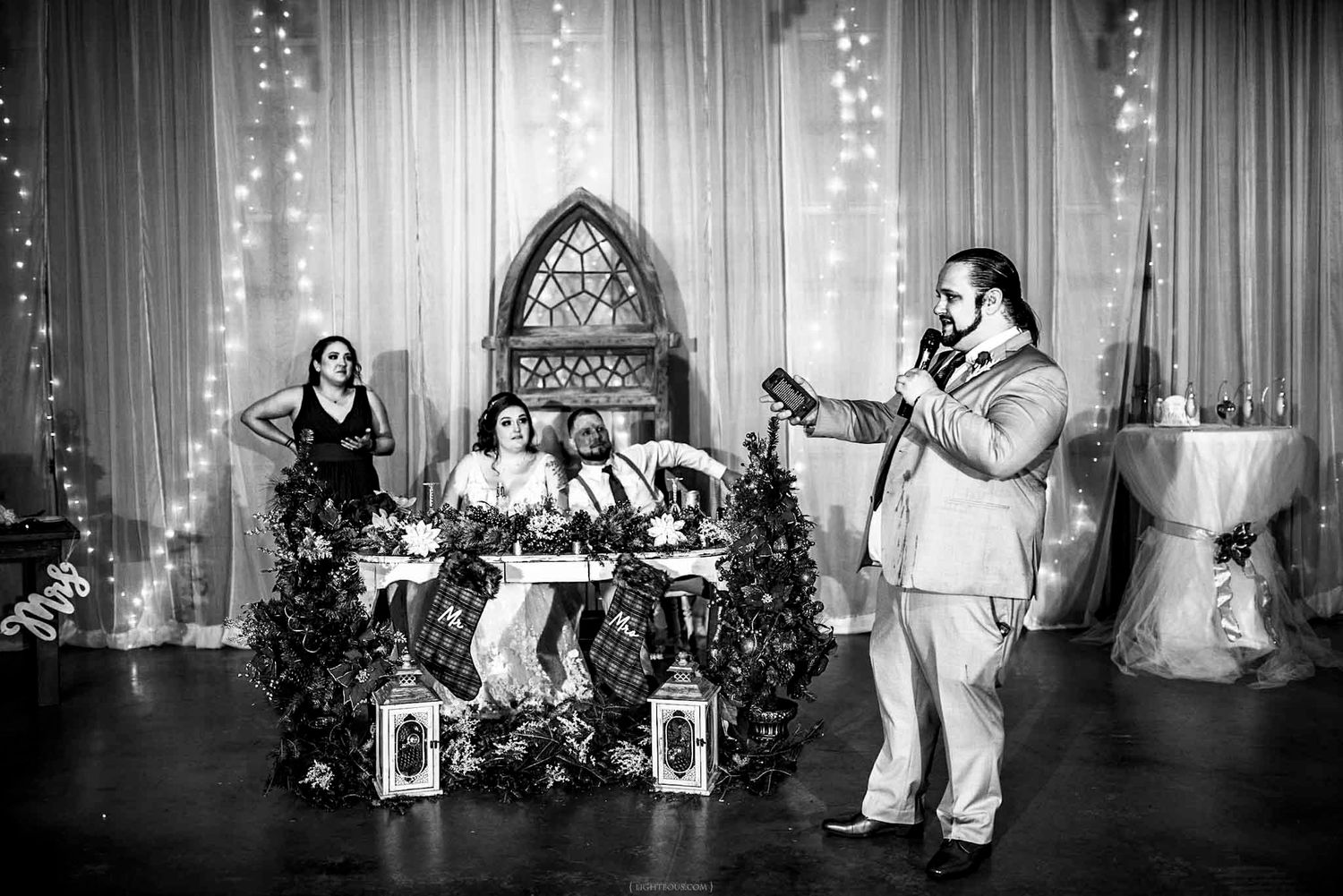 Black and white wedding reception photo with groom giving a toast at decorated sweetheart table with fairy lights.