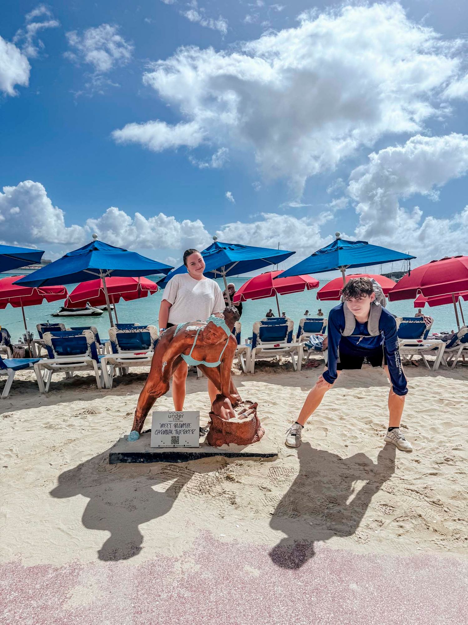 Two tourists pose with a decorative horse statue on a sunny Caribbean beach with colorful umbrellas.