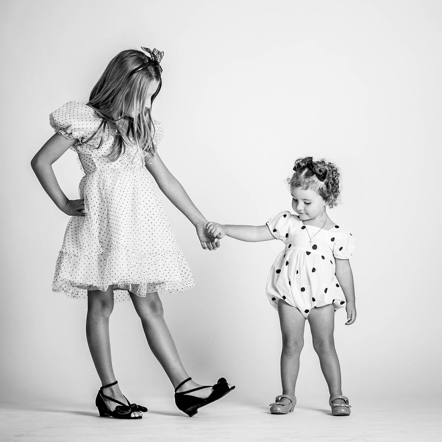 Two young sisters hold hands in a black and white photo, older girl in polka dot dress, toddler in romper.