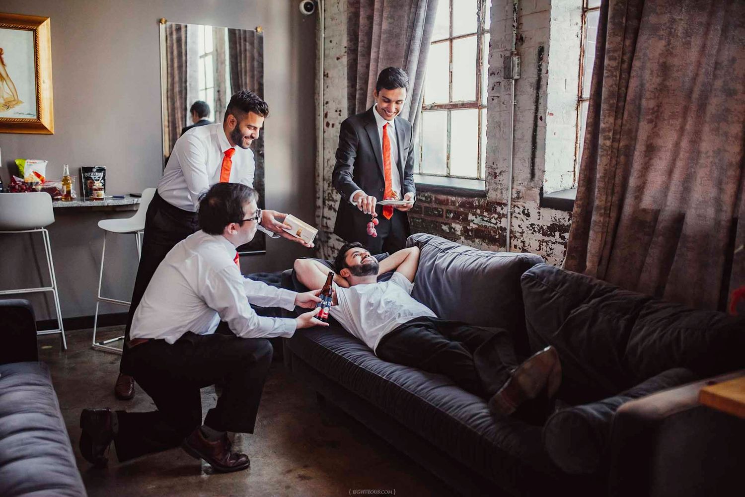 Groomsmen in dress shirts and ties helping each other prepare on a couch in a rustic wedding venue.