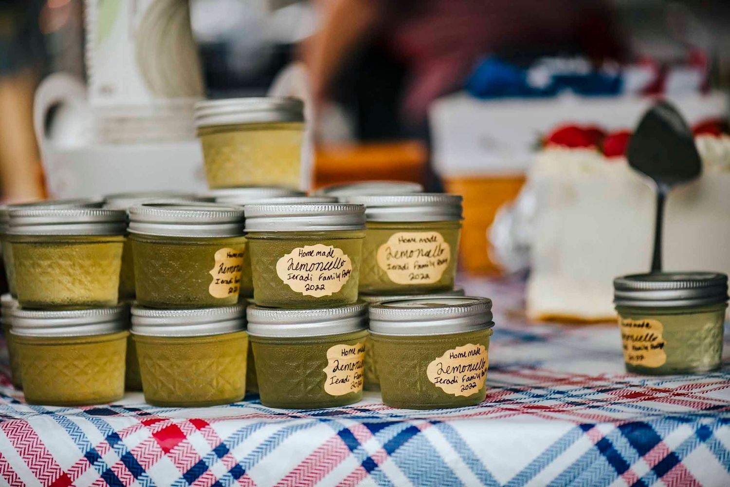Homemade jams and preserves in mason jars displayed on a plaid tablecloth at an outdoor farmers market.