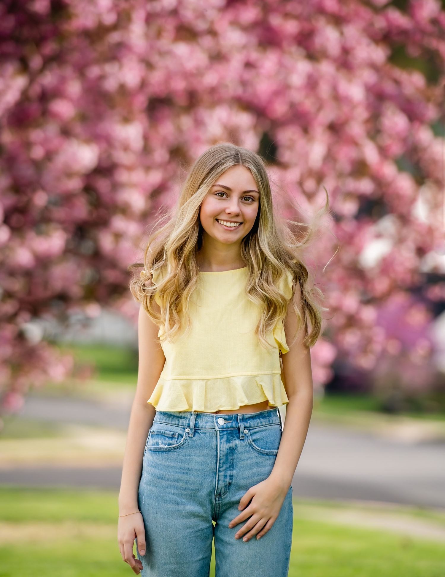 Young blonde woman in yellow ruffle crop top and jeans smiling outdoors with pink cherry blossom tree background.