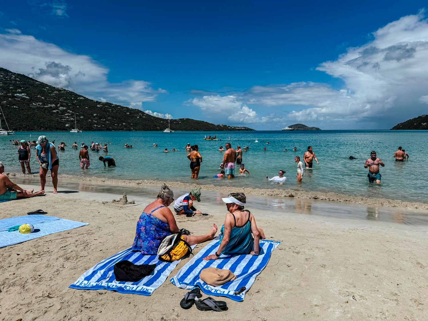 Tourists relaxing on blue striped towels on a sandy Caribbean beach with calm turquoise waters and green hills.