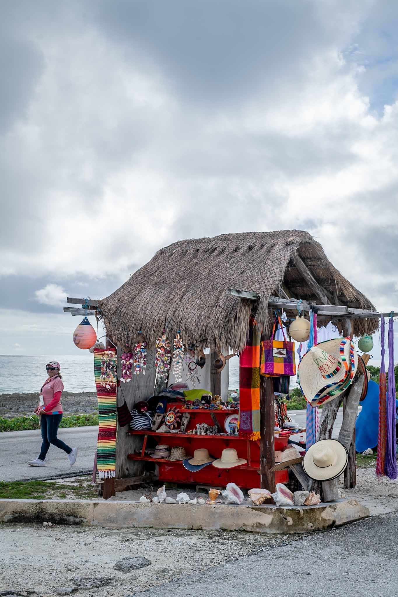 Row of thatched-roof souvenir shops selling colorful merchandise along a beachfront in Cozumel, Mexico.