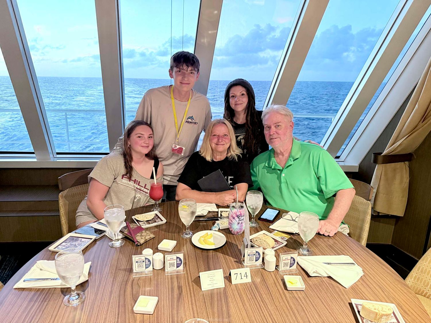 Family of five smiling together at a dining table on a cruise ship with ocean views through large windows.