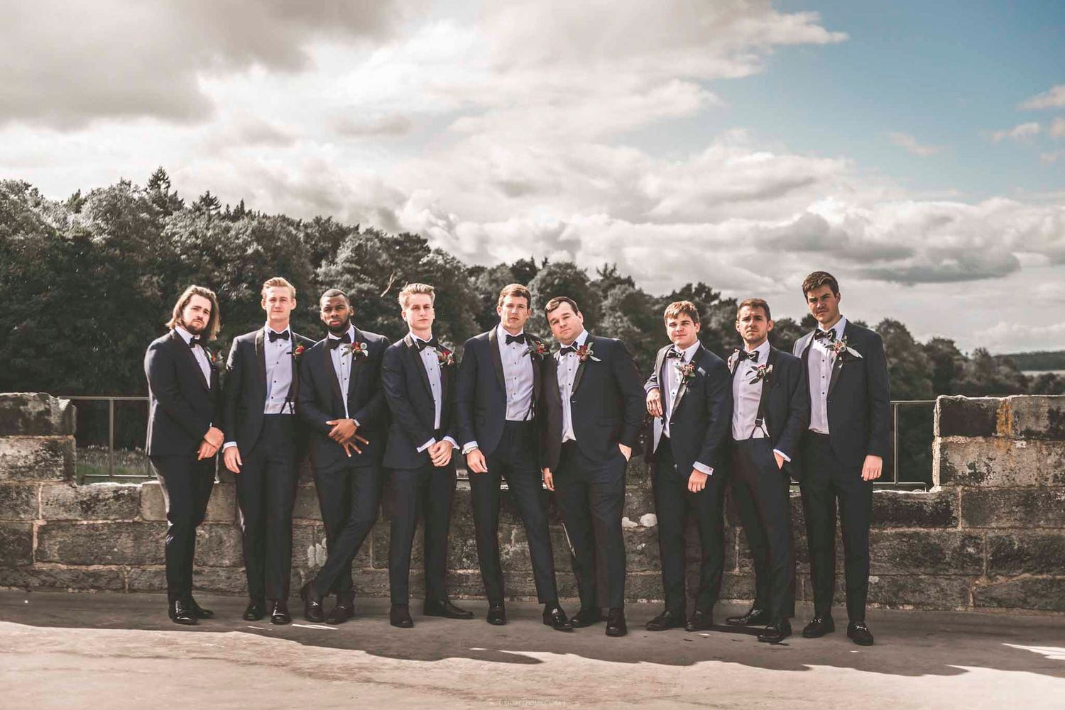 Groomsmen in black tuxedos and bow ties posing outdoors on a stone terrace under a cloudy sky.
