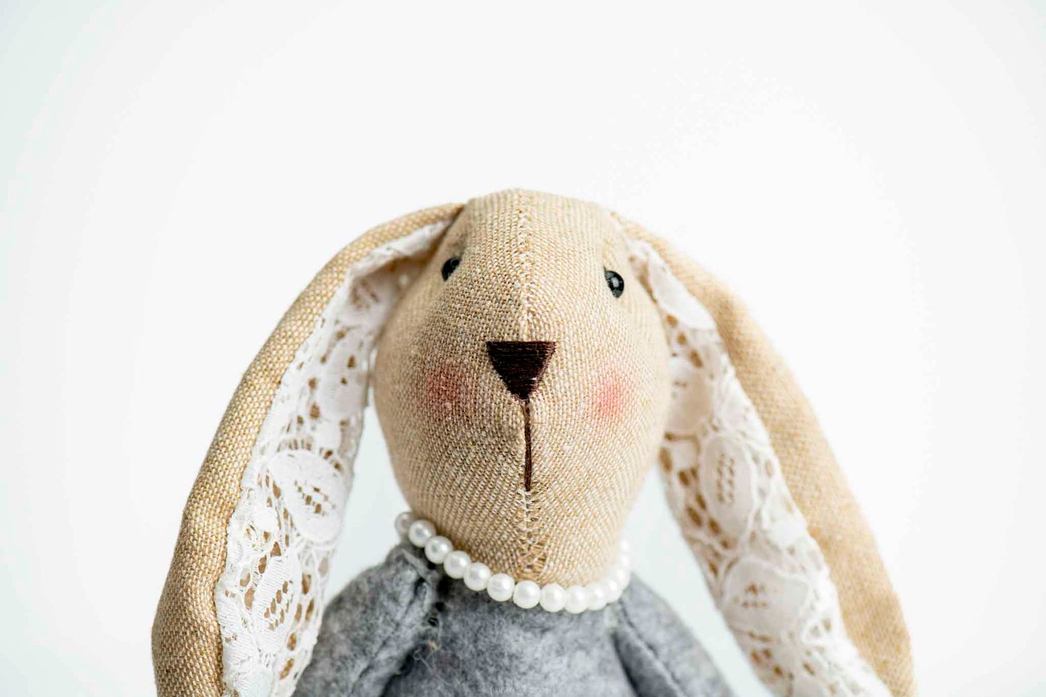 Handmade fabric bunny toy with lace ears, pearl necklace, and rosy cheeks on white background.