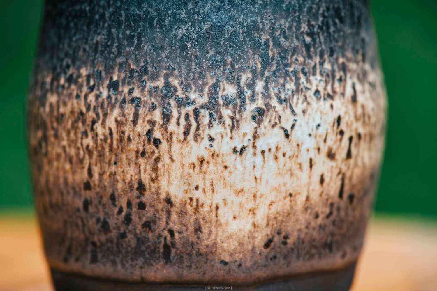 Close-up of a rustic ceramic vase with textured brown and gray glaze, set against a green background.