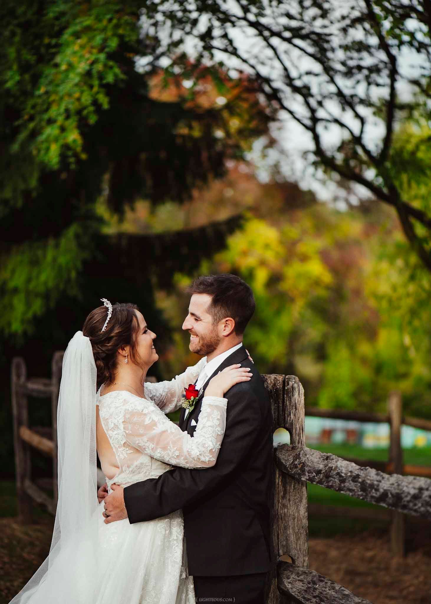 Bride and groom share a romantic moment by a rustic fence surrounded by lush autumn greenery.
