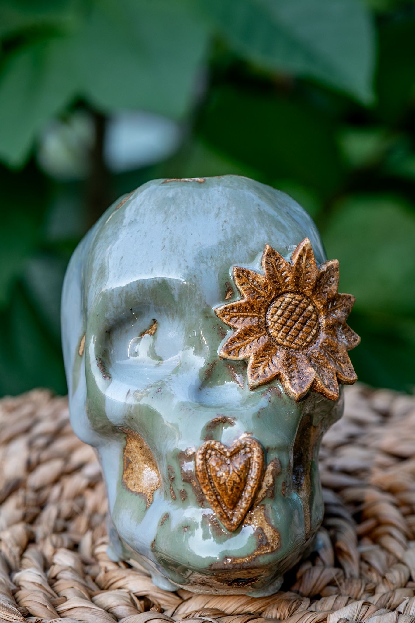 Ceramic skull with golden sunflower and heart decorations in teal glaze on wicker surface.