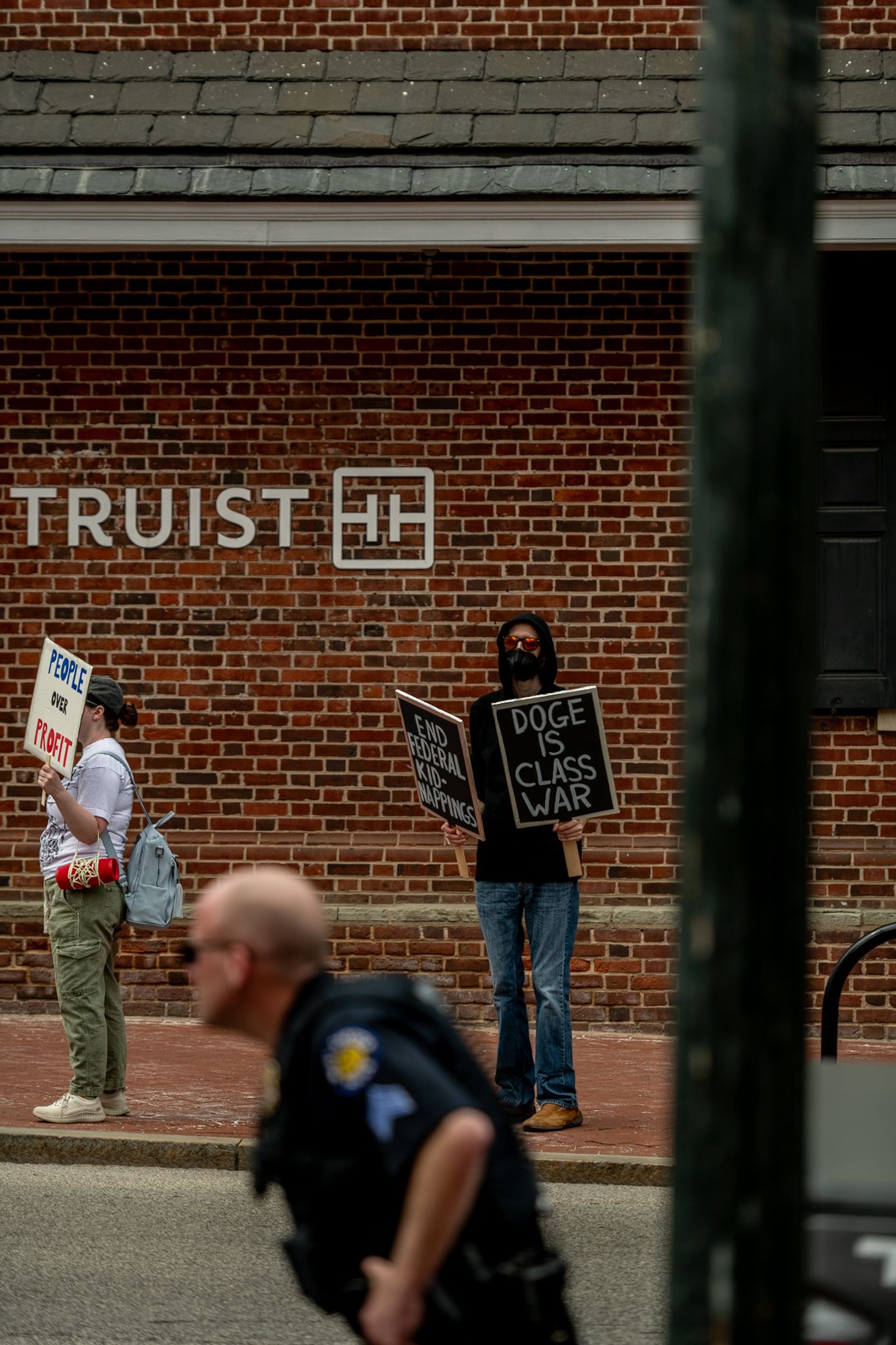 Protesters with signs gathered near brick wall during demonstration.