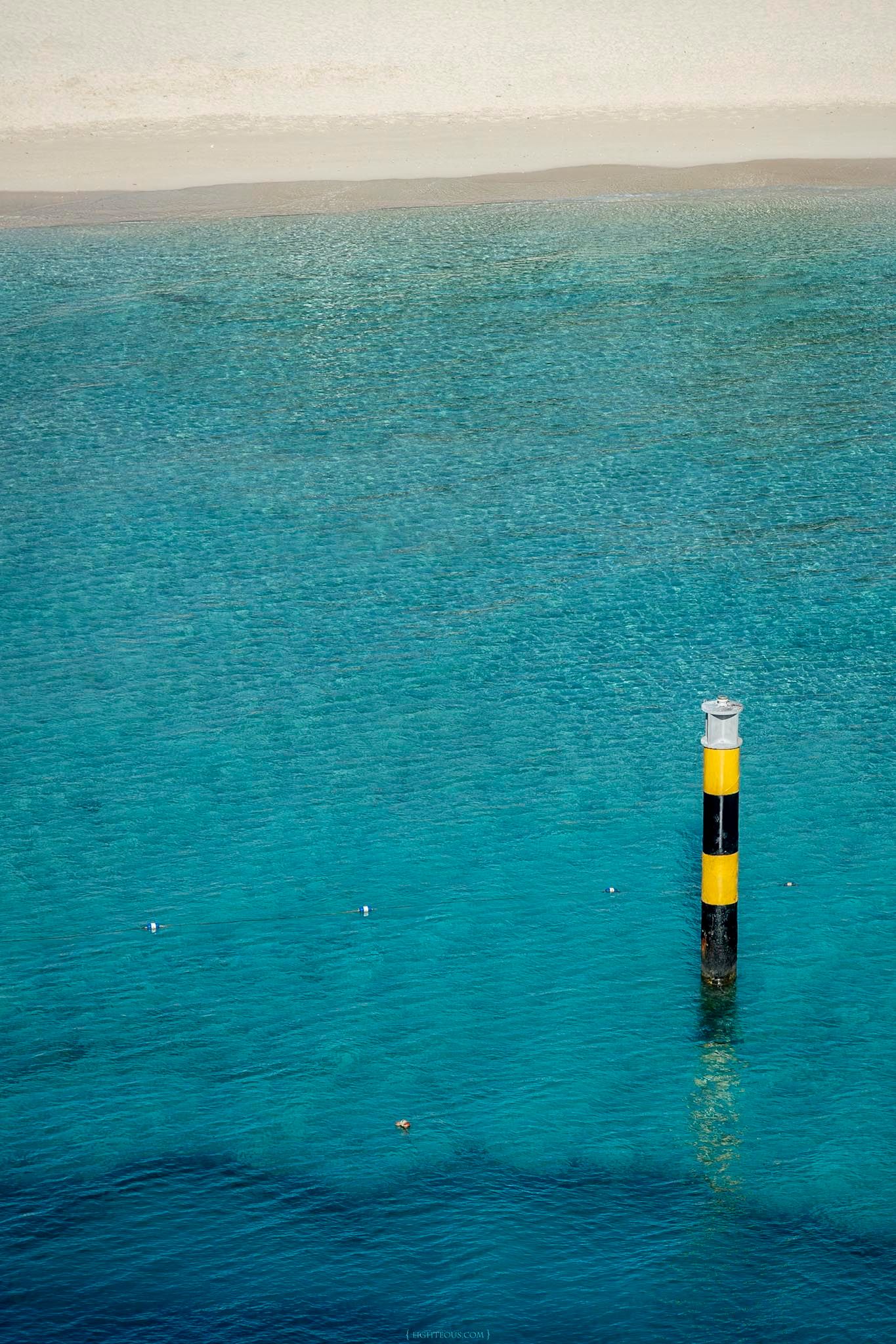 Aerial view of a yellow and black striped navigation marker pole standing in clear turquoise ocean water near a sandy beach.