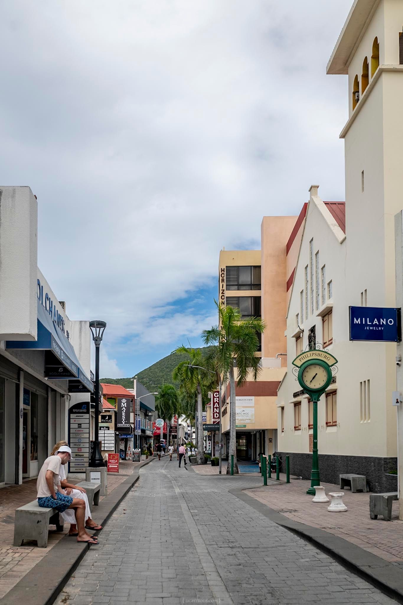 Pedestrian street in Philipsburg, Sint Maarten with Milano Jewelry store, street clock, and mountain backdrop.