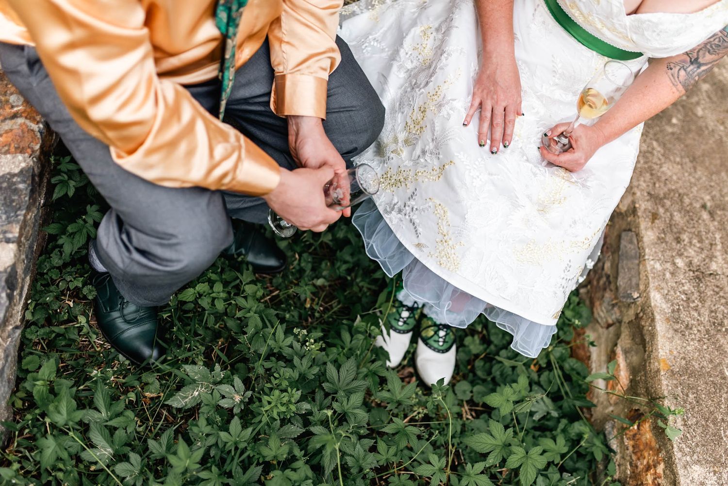 Bride and groom holding hands while sitting outdoors surrounded by lush green ground cover on their wedding day.