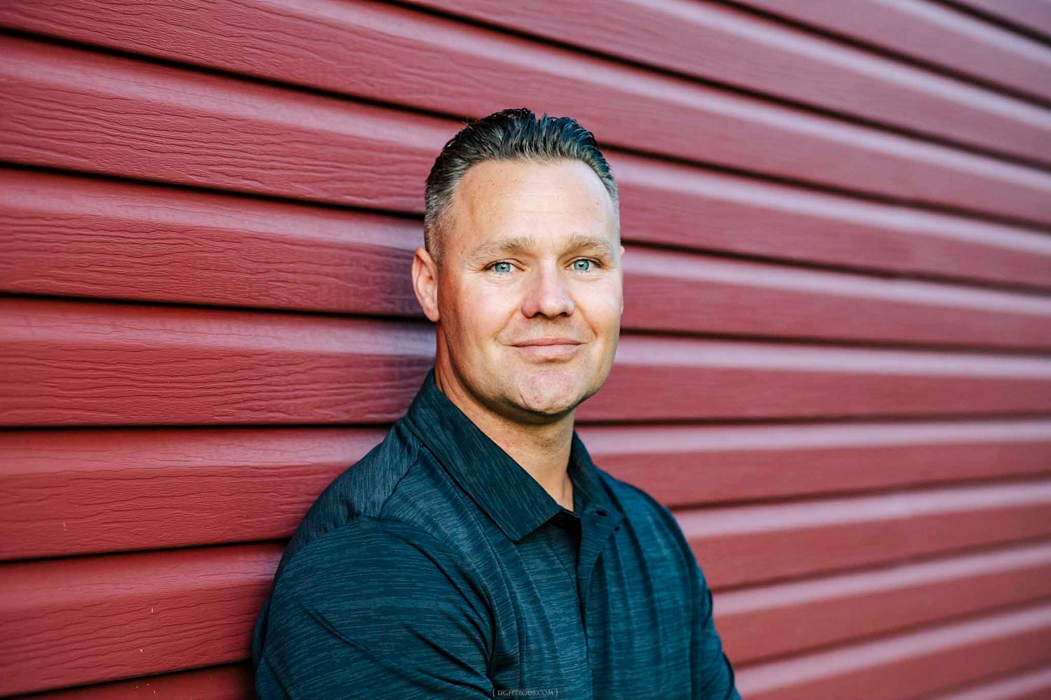 Smiling man in dark shirt posing against red wooden siding wall outdoors in natural light.