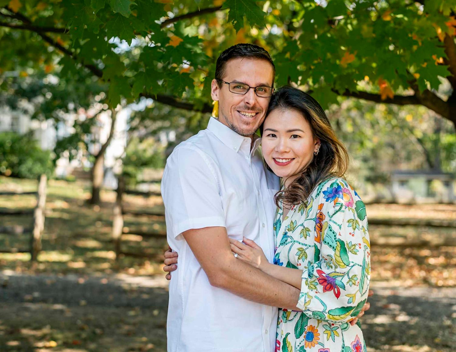 Couple embracing outdoors under green trees, man in white shirt and woman in floral dress, smiling portrait photo.