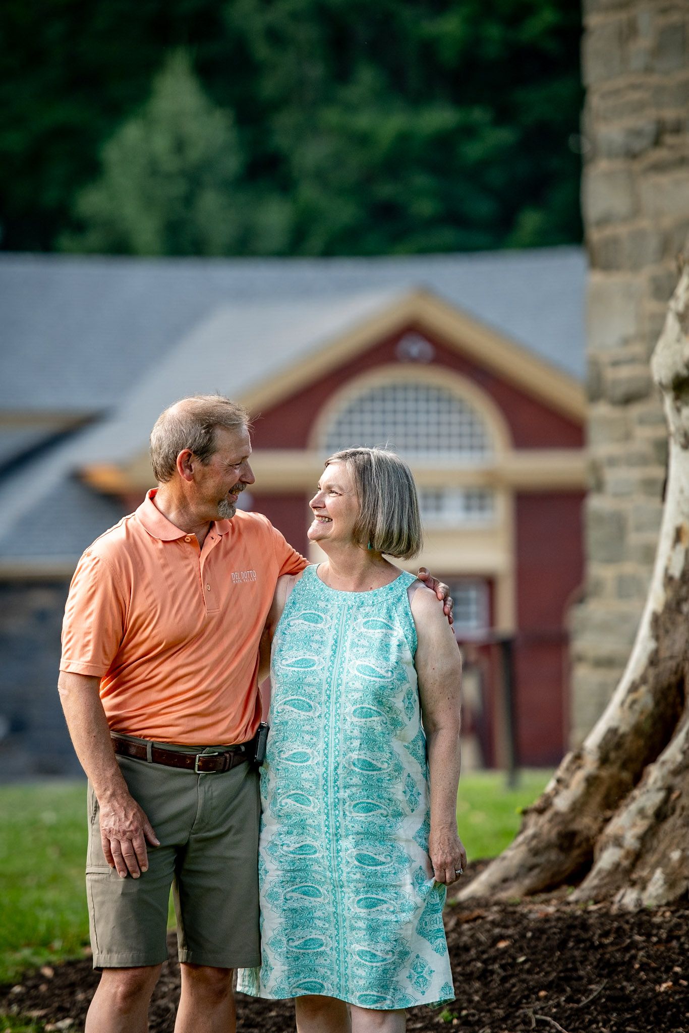 A smiling older couple stands together outdoors near a large tree, with a red brick building visible in the background.