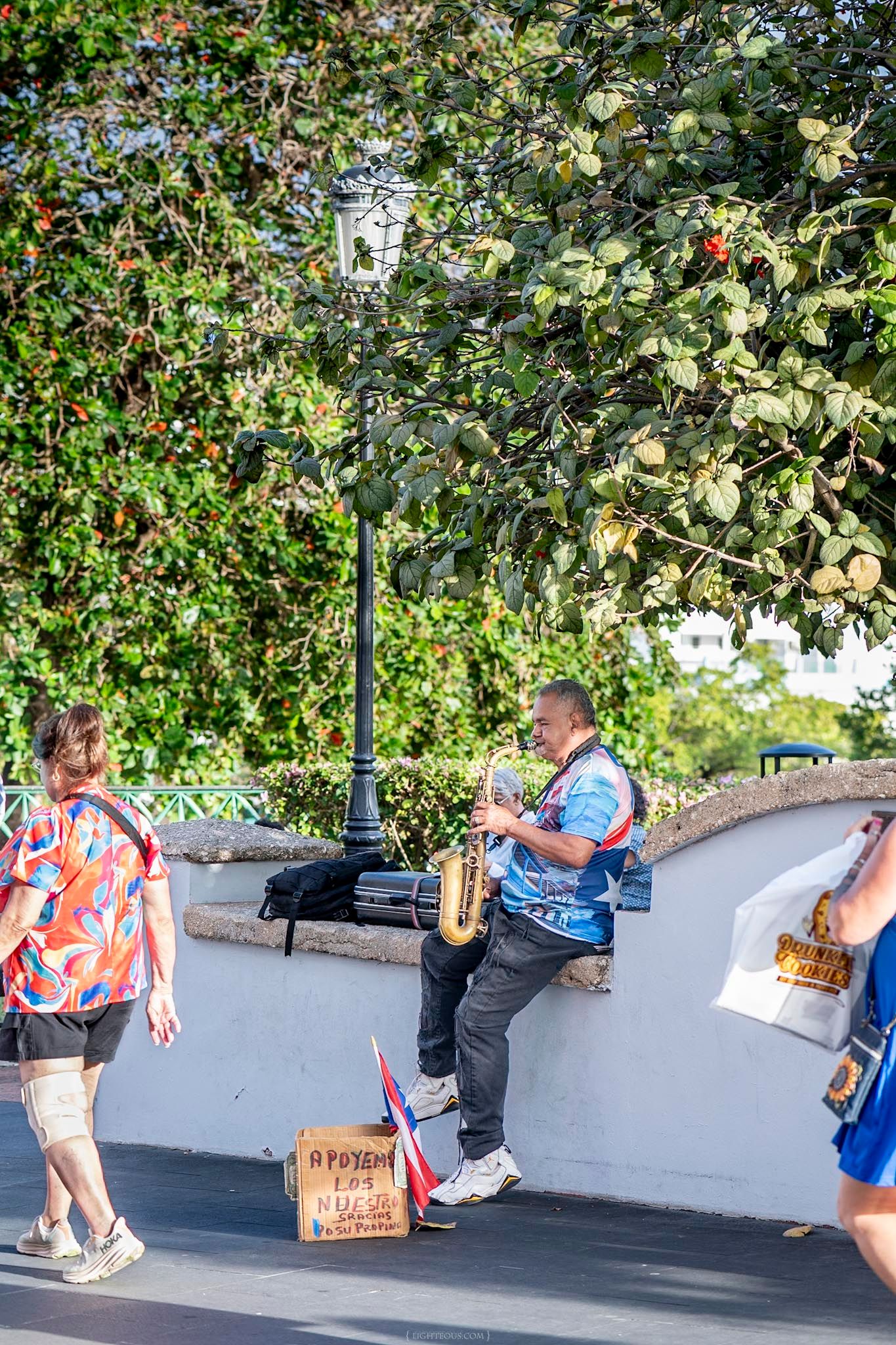 Street musician plays saxophone near a lamp post in Old San Juan, Puerto Rico, with lush green trees in the background.